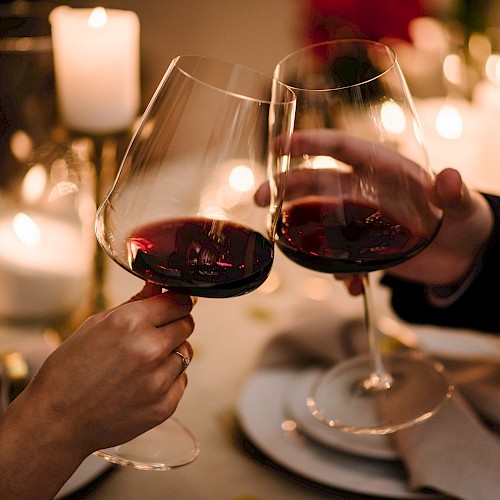 Two people clinking glasses of red wine at a candlelit table, surrounded by cozy, warm lighting and an elegant dinner setting.