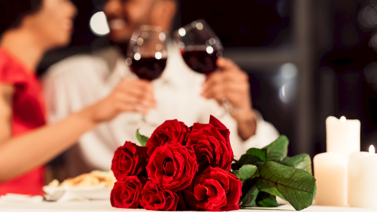 A couple is toasting with red wine glasses at a candlelit dinner, with a bouquet of red roses in the foreground.