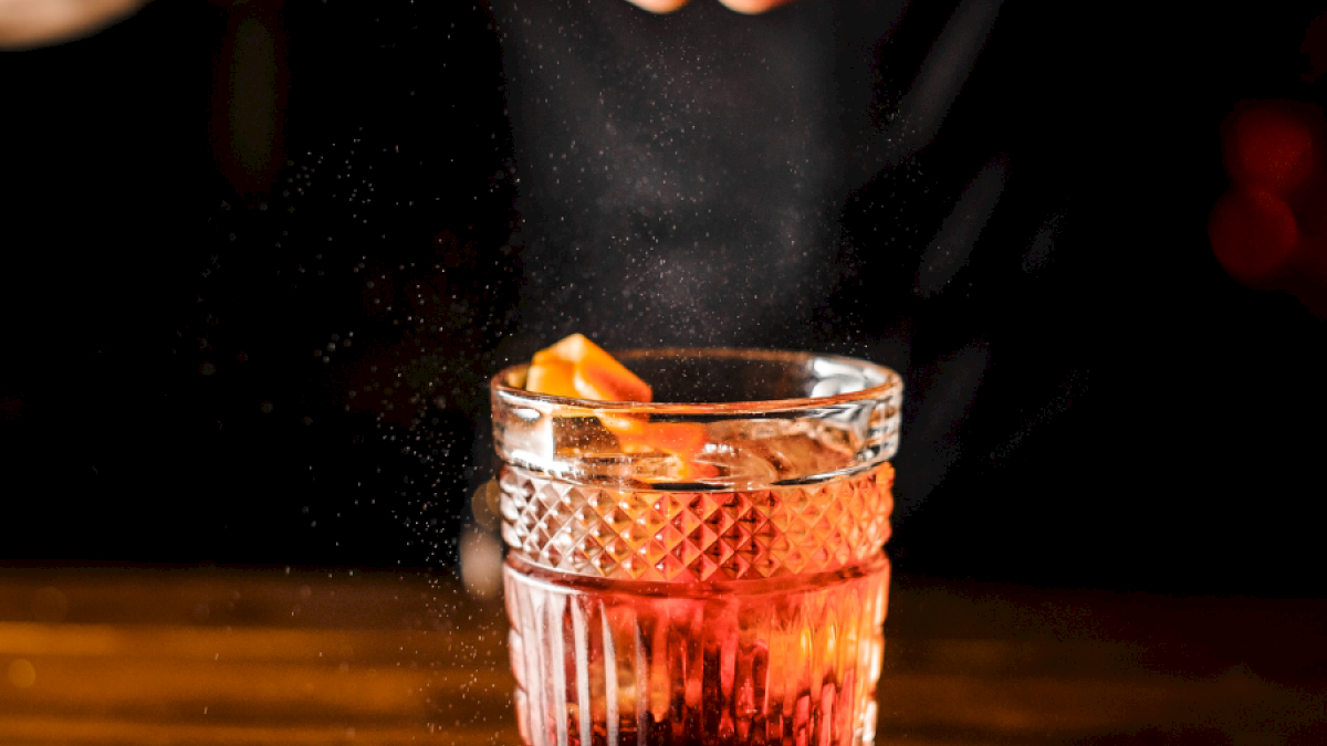 Someone is adding a citrus peel to a glass of dark red cocktail, garnished with a citrus slice, on a dark table.