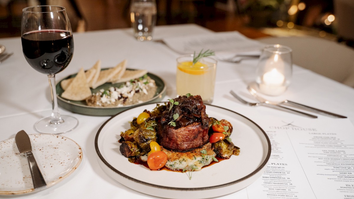 A beautifully plated main course with steak, Brussels sprouts, cherry tomatoes, and mashed potatoes, alongside a glass of red wine.