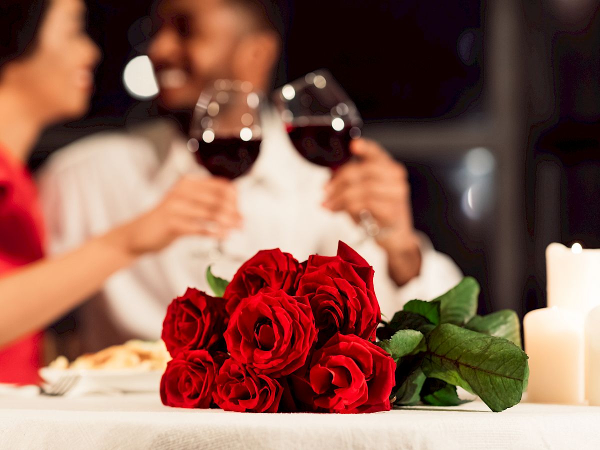 A couple sits at a candlelit table with red roses in the foreground, sharing a toast with glasses of red wine.