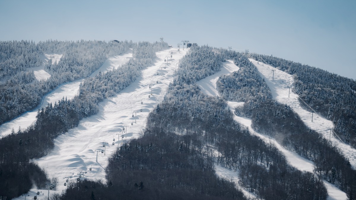 This image depicts a snow-covered mountain with ski slopes and chairlifts. Trees are partially covered in snow, and the sky is clear blue.