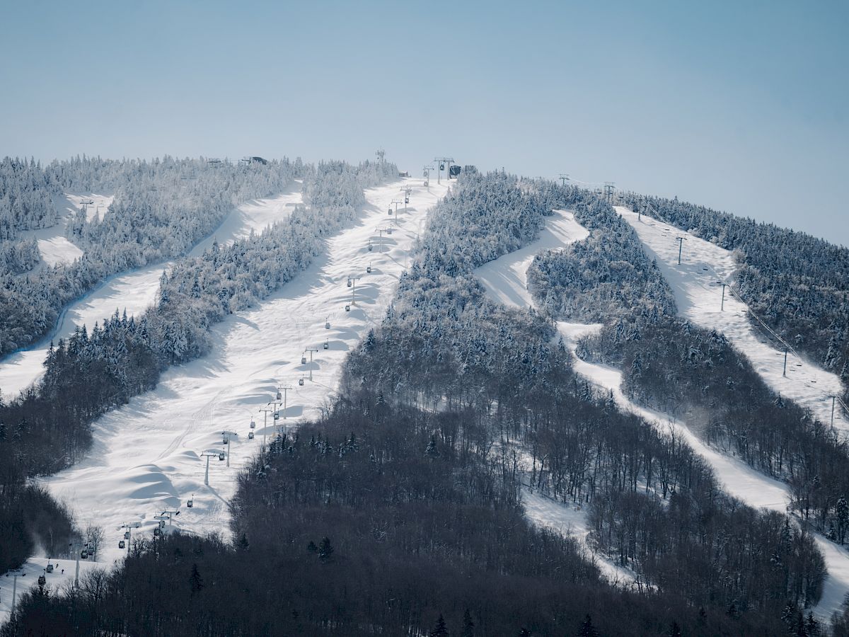 This image depicts a snow-covered mountain with ski slopes and chairlifts. Trees are partially covered in snow, and the sky is clear blue.