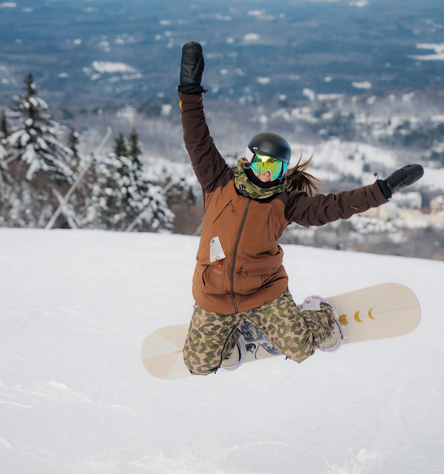 A snowboarder wearing a helmet and goggles is kneeling in the snow with arms raised in a victory pose, surrounded by a snowy mountain landscape.