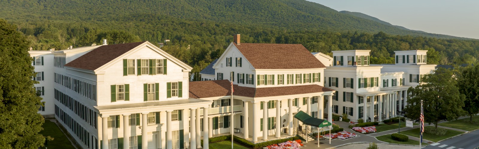 A large white hotel with green shutters, manicured gardens, and a mountain backdrop under a clear sky.