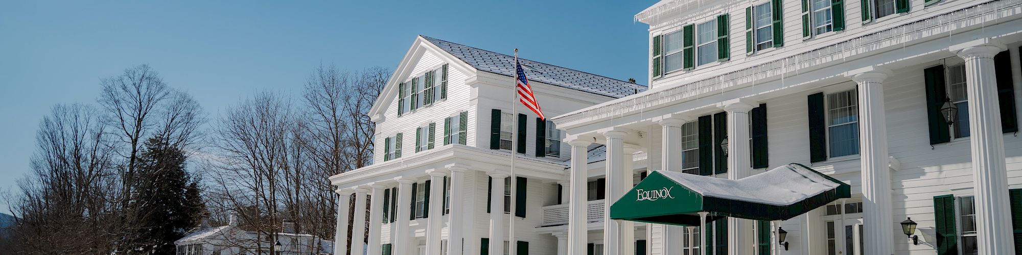 A snowy street with a large, white building featuring green shutters and an American flag, along with a green awning. Trees are in the background.