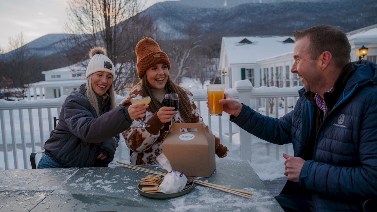 Three people are enjoying drinks and celebrating outdoors in a snowy setting, with mountains in the background and warm clothing on them.