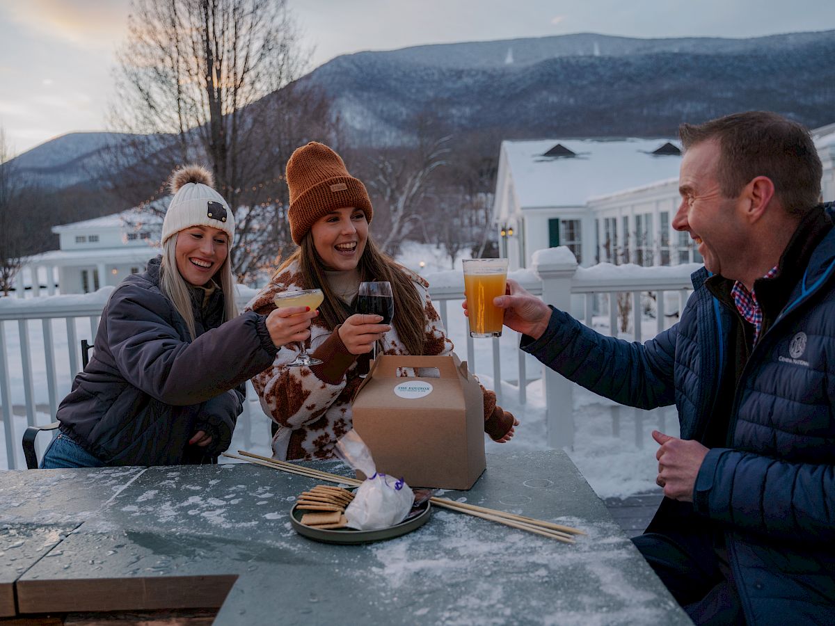 Three people are enjoying drinks and celebrating outdoors in a snowy setting, with mountains in the background and warm clothing on them.