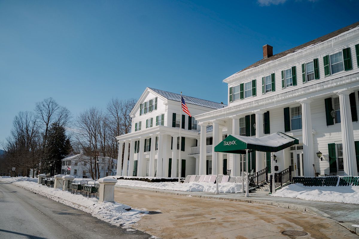 An image of a snowy street with large white buildings featuring many windows and columns, and an American flag displayed outside.