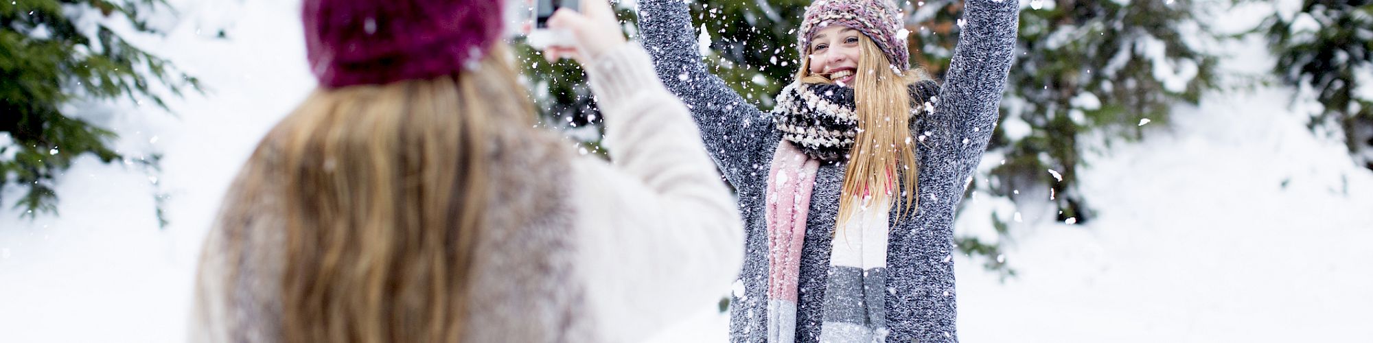 Two people are in a snowy forest; one is taking a photo of the other who is joyfully playing in the snow with arms raised.