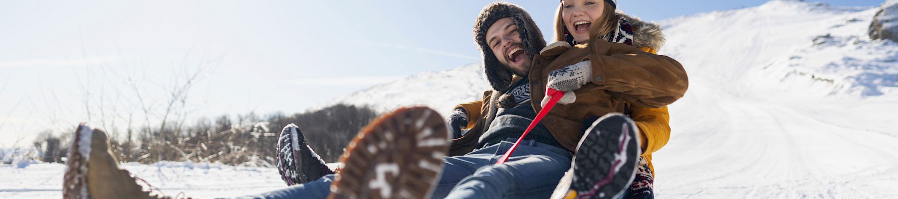 Two people are sledding down a snowy slope, laughing and enjoying the ride on a bright, sunny day.