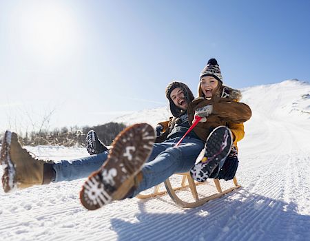 Two people are sledding down a snowy slope, laughing and enjoying the ride on a bright, sunny day.