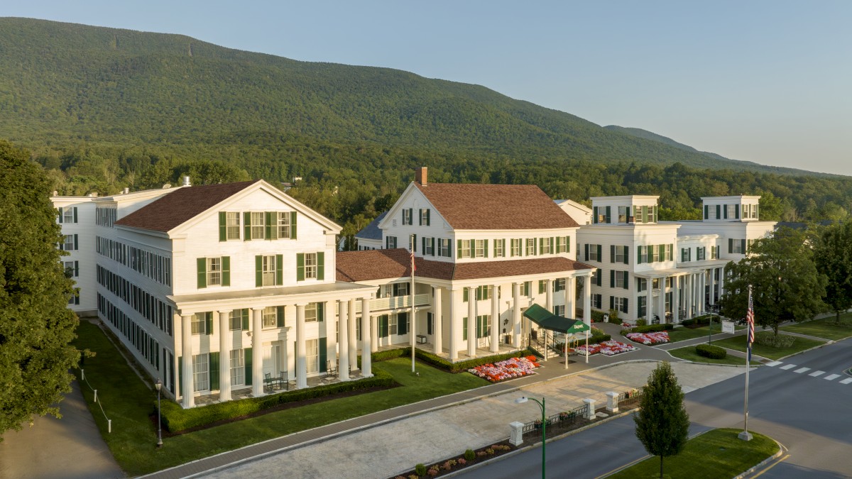 An aerial view of a large, white, colonial-style building with green shutters, situated against a backdrop of forested hills.