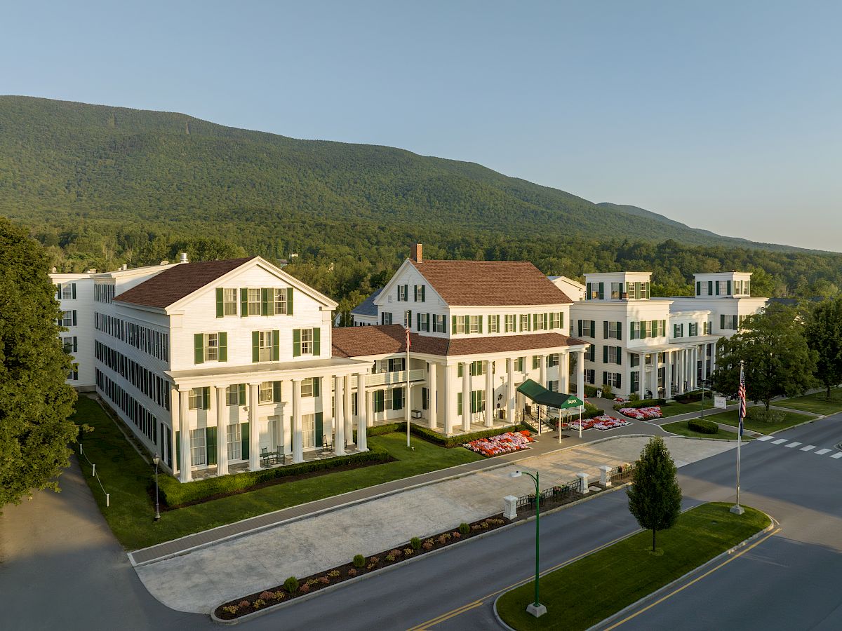 An aerial view of a large, white, colonial-style building with green shutters, situated against a backdrop of forested hills.