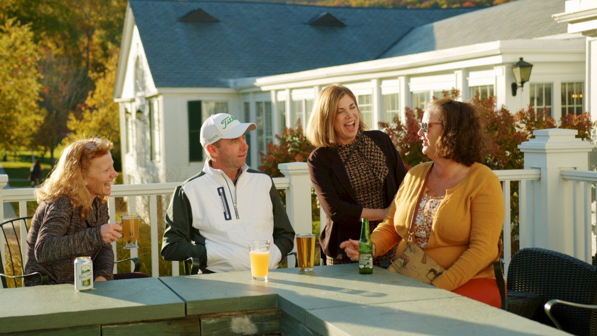 A group of four people, two men and two women, are sitting at an outdoor table, laughing and enjoying drinks on a sunny day.