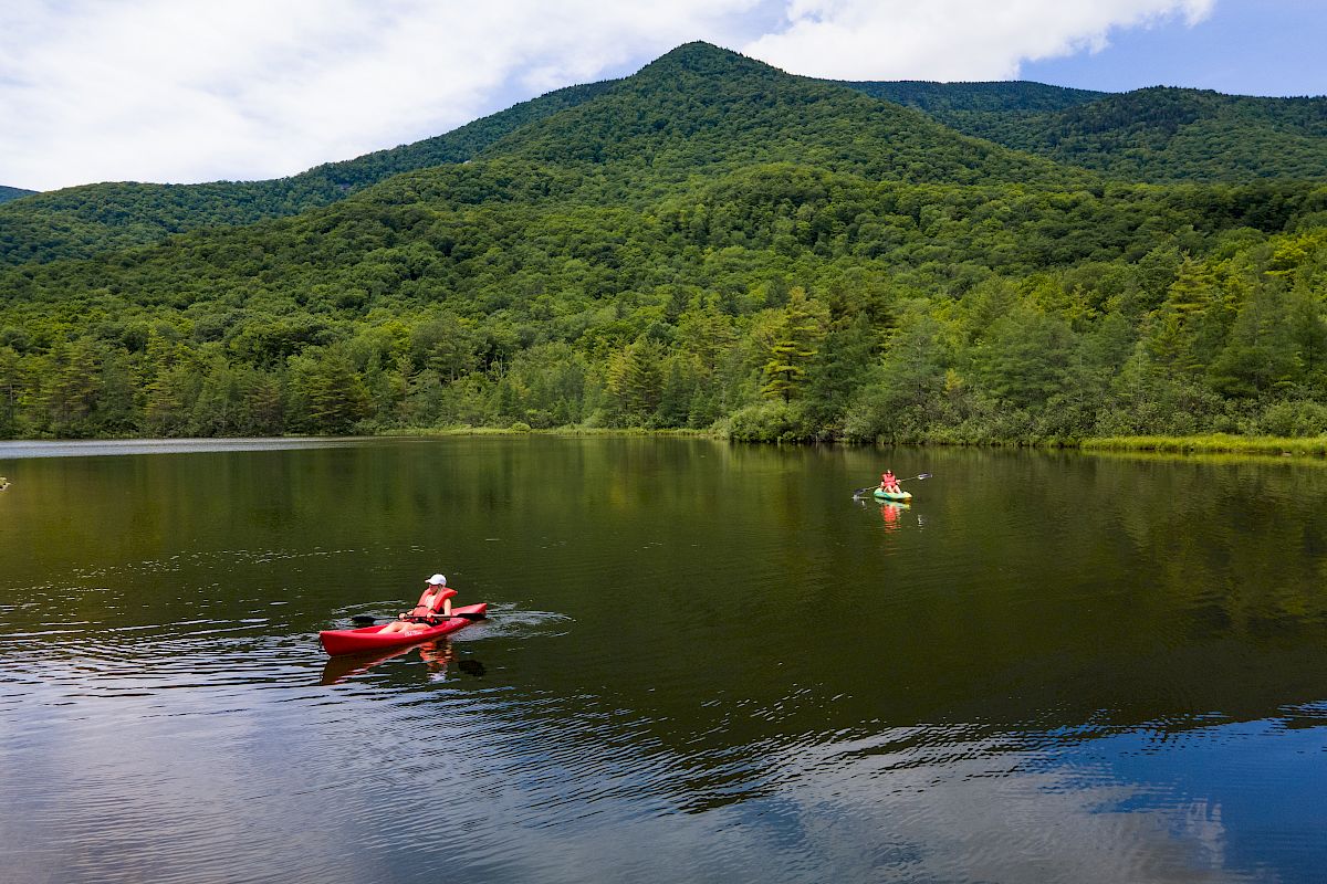 Two people kayaking on a calm lake surrounded by lush green forested hills under a partly cloudy sky.