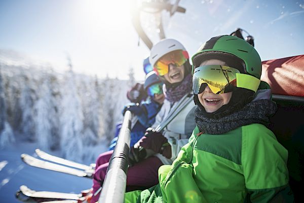 A group of people in winter gear, including helmets and goggles, ride a ski lift with snow-covered trees in the background under a bright sky.