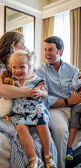 A family of four is sitting on a bed in a cozy room, smiling and enjoying time together, with toys and books.