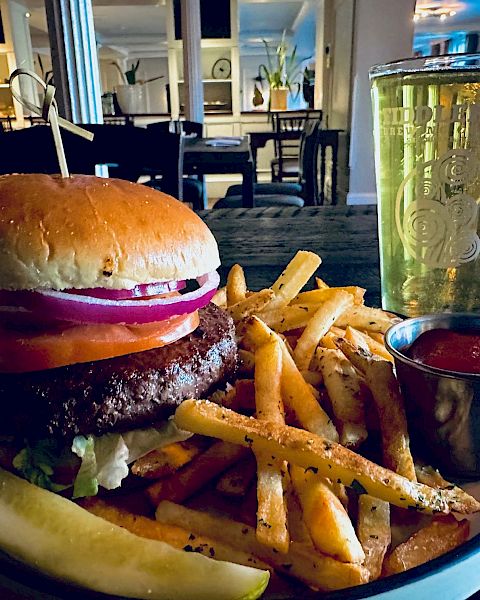A classic burger with lettuce, tomato, onion, fries, a pickle spear, ketchup on the side, and a glass of beer on a table in a cozy restaurant setting.