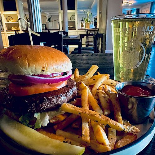 A classic burger with lettuce, tomato, onion, fries, a pickle spear, ketchup on the side, and a glass of beer on a table in a cozy restaurant setting.