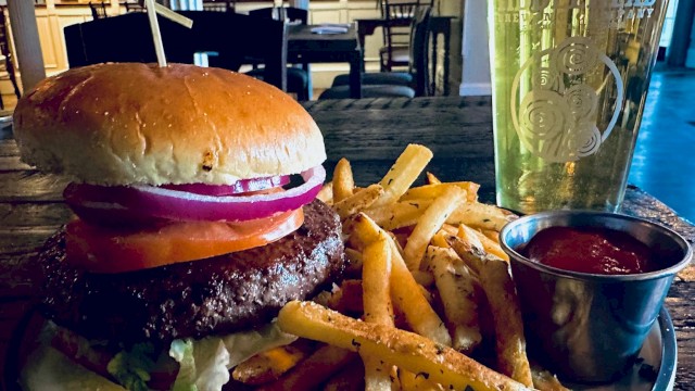 A classic burger with lettuce, tomato, onion, fries, a pickle spear, ketchup on the side, and a glass of beer on a table in a cozy restaurant setting.