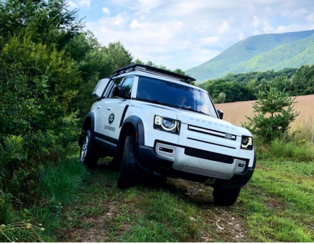 A white Land Rover SUV is off-roading on a green trail with mountains and a partly cloudy sky in the background.