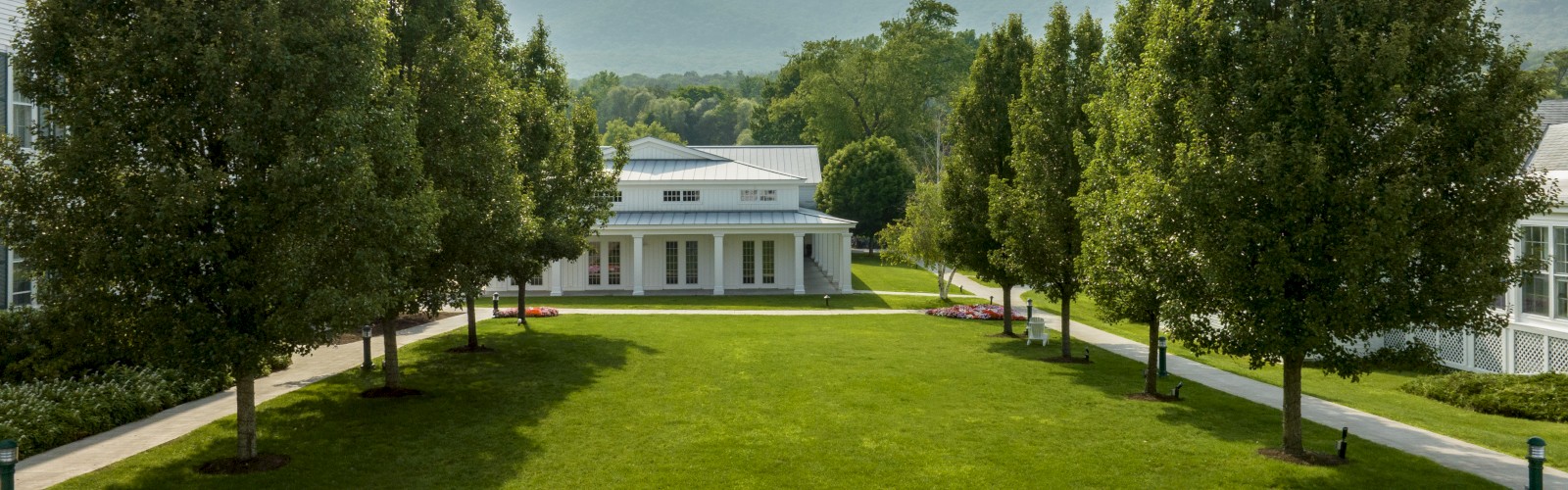 A well-manicured lawn with a path lined by trees, a small building in the center, and mountains in the background under a clear sky.
