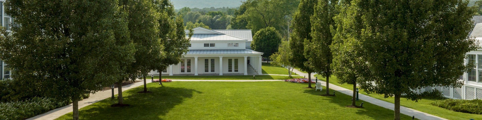 A well-manicured lawn with a path lined by trees, a small building in the center, and mountains in the background under a clear sky.