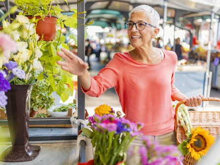 A person with short white hair and glasses is smiling while selecting flowers at a market; holding a basket with sunflowers.