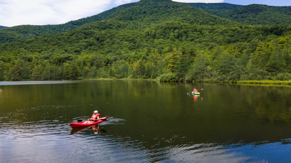 Two people are kayaking on a calm lake surrounded by green hills and mountains under a partly cloudy sky.