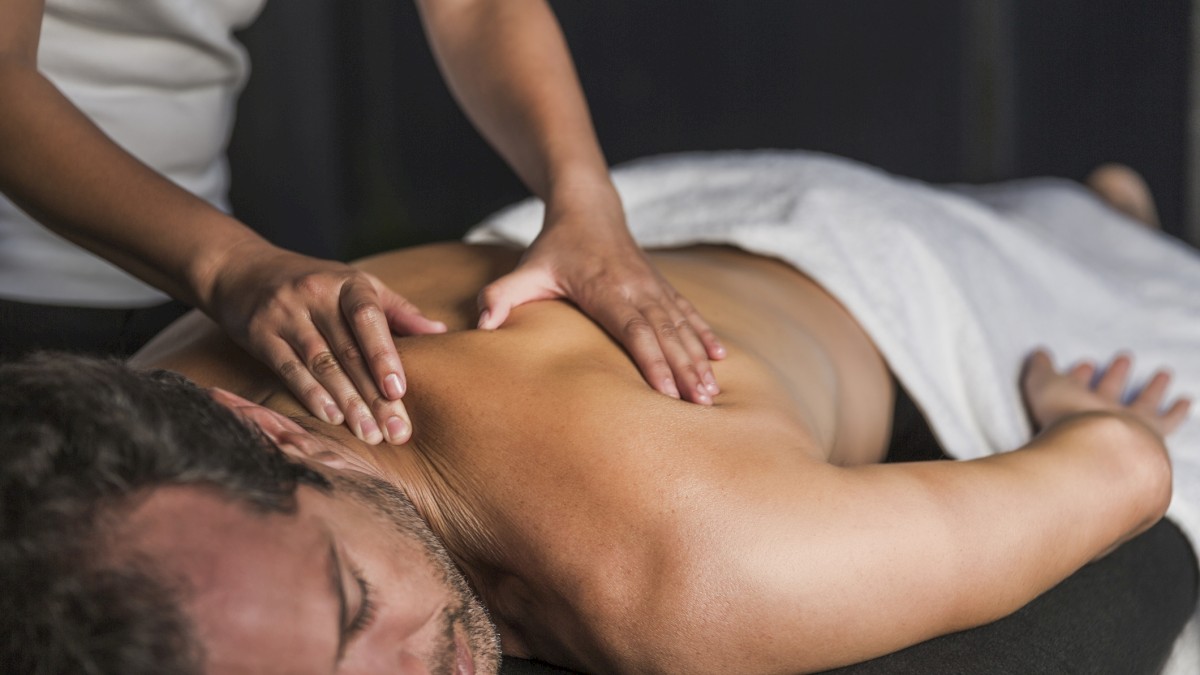 A person is laying on a massage table, covered partially by a towel, and receiving a back massage from a therapist.