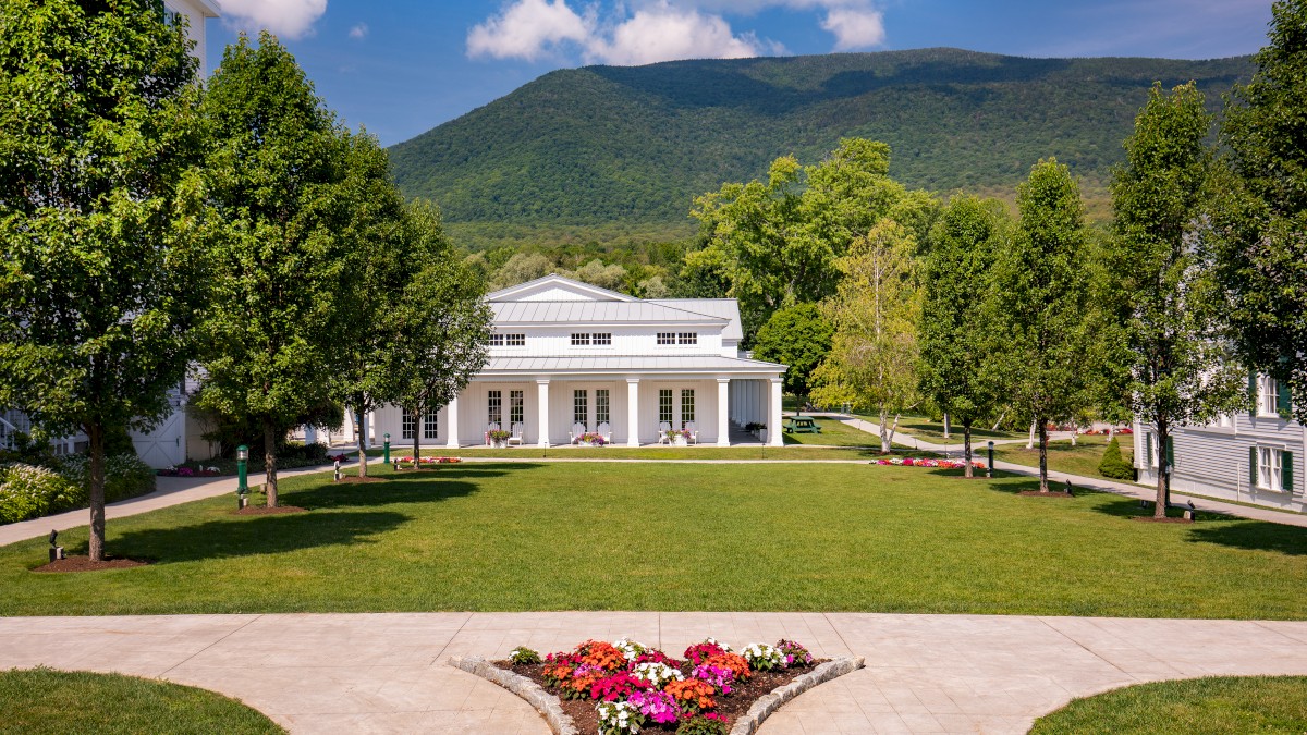 A scenic view featuring a white building with columns, surrounded by lush green trees, a manicured lawn, and mountains in the background.