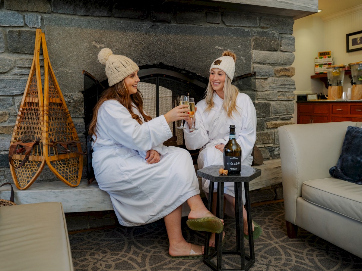 Two people in robes and beanies toast drinks in front of a fireplace, with snow shoes on the wall and a bottle on a small table.