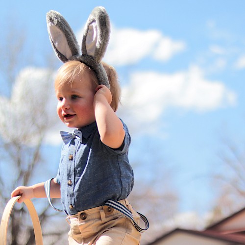 A child wearing bunny ears holds a basket outside under a clear sky with trees and buildings in the background.