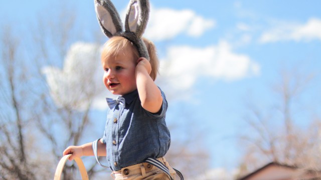 A child wearing bunny ears holds a basket outside under a clear sky with trees and buildings in the background.