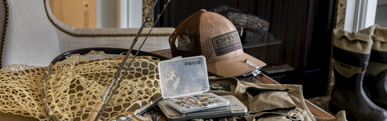 Fishing gear is displayed on a table, including a fishing rod, hat, tackle box, flies, net, and vest, with boots visible near a fireplace.