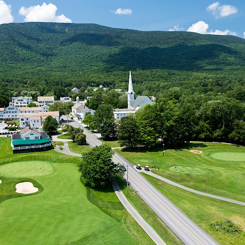 An aerial view of a town with a white church, surrounded by lush greenery, golf course, and mountains. The sky is partly cloudy.