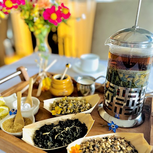 A tray with a French press of tea, lemon slices, honey, various loose teas, a sugar bowl, and a small vase of flowers on a table.