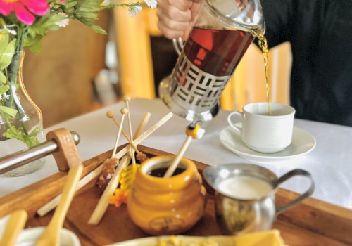 A person pouring tea from a French press into a cup, with a tray containing honey, milk, flowers, and other tea ingredients, is in the image.
