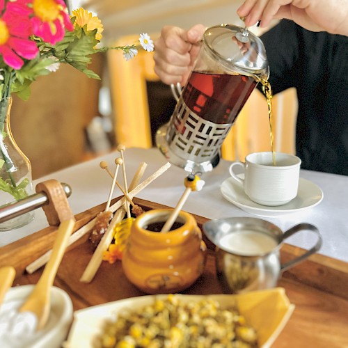 A person pouring tea from a French press into a cup, with a tray containing honey, milk, flowers, and other tea ingredients, is in the image.