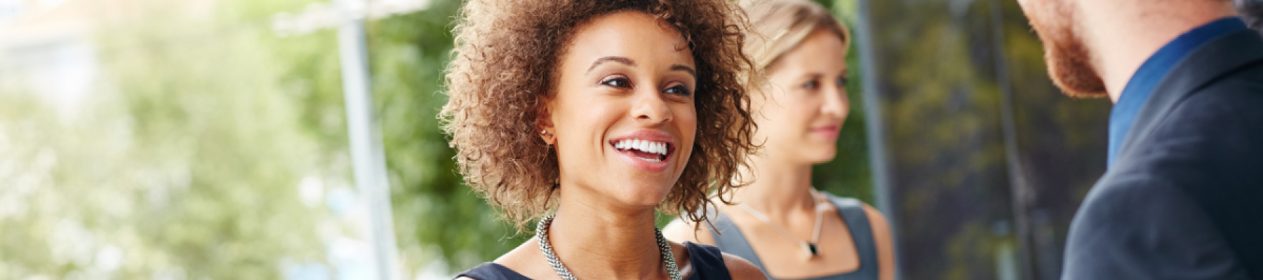 A woman with curly hair, wearing a black dress, is smiling and shaking hands with a man. A woman in the background is holding a folder.