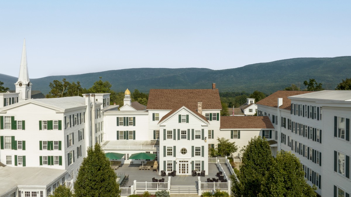 This image shows a large, elegant white building complex with green shutters and a central courtyard, set against a backdrop of hills and clear skies.