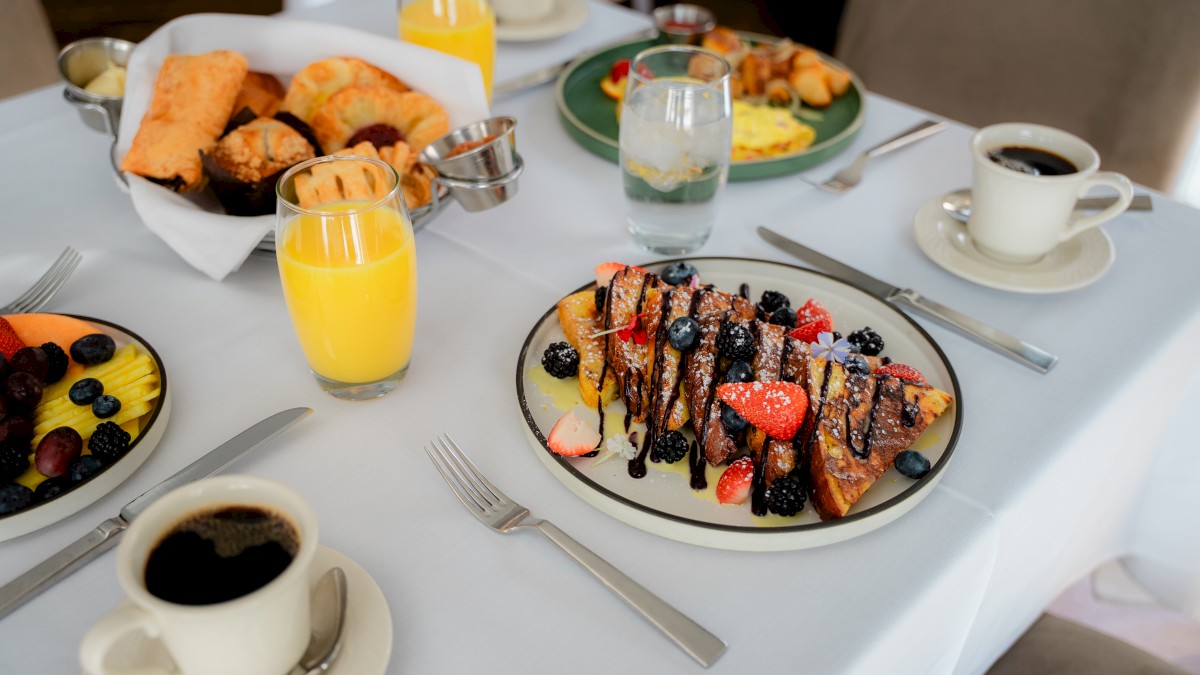 A breakfast spread with fruit, French toast with berries, pastries, orange juice, water, and coffee on a white tablecloth.