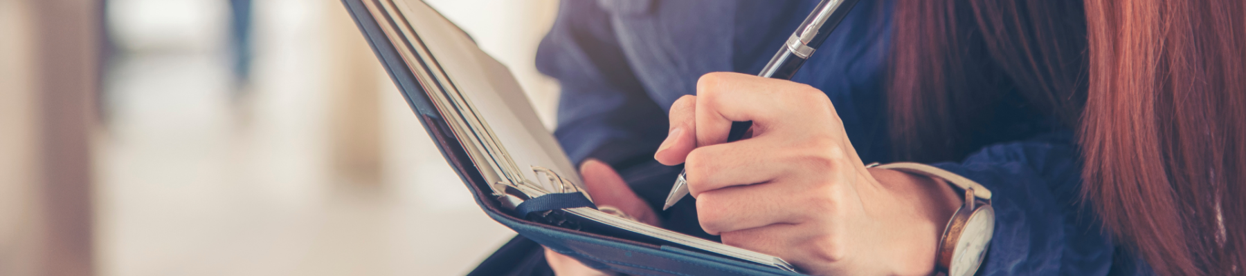 A person is multitasking, holding a phone between their ear and shoulder while writing in a notebook with a pen in hand, dressed in blue attire.