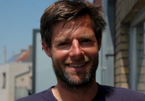 A man with short hair and a beard is smiling outside in a sunny area with buildings in the background.