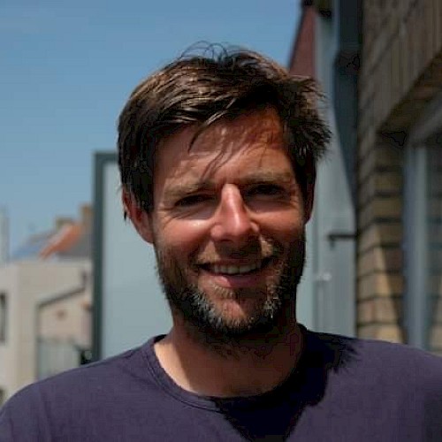 A man with short hair and a beard is smiling outside in a sunny area with buildings in the background.