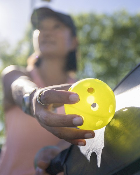 A person holding a yellow perforated ball and a paddle, likely used in pickleball. The person is outdoors with greenery in the background.