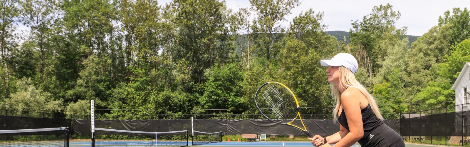 A person in athletic gear holds a tennis racket while preparing to play on an outdoor tennis court surrounded by trees.