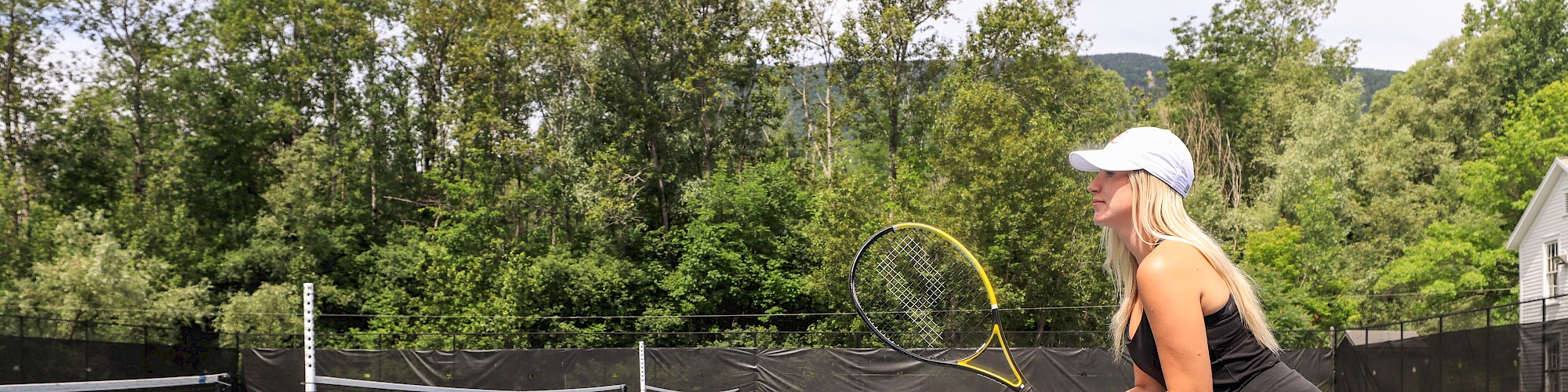 A person in athletic gear holds a tennis racket while preparing to play on an outdoor tennis court surrounded by trees.