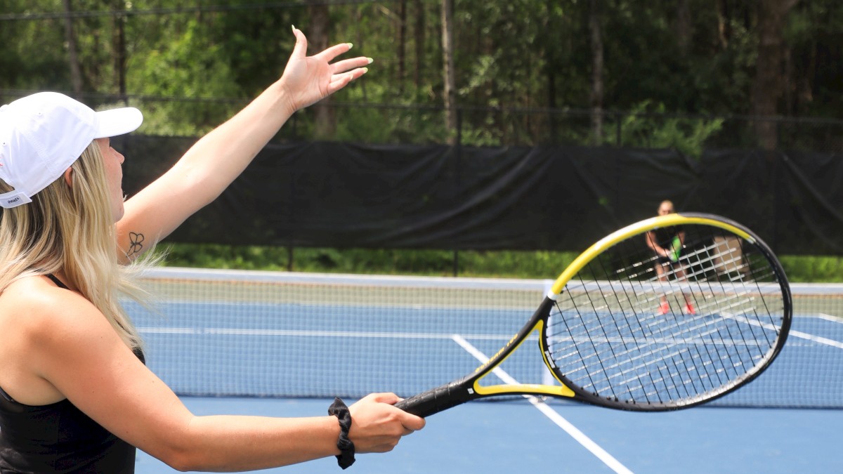 A person is serving a tennis ball on a blue outdoor court, with trees in the background.